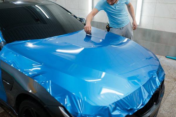 Car wrapping, man with squeegee installs protective vinyl foil or film on hood. Worker makes auto detailing. Automobile paint protection coating, professional tuning